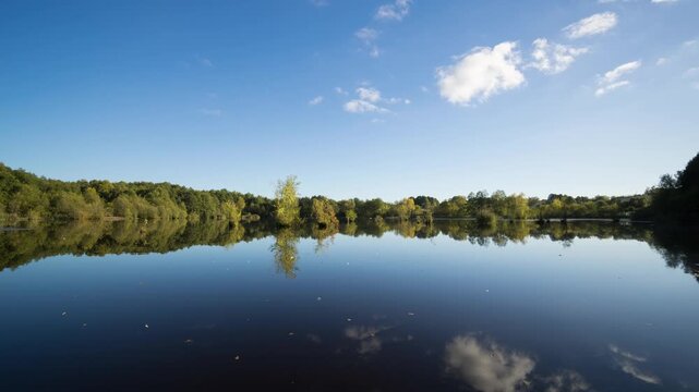 Time lapse of sunny autumn lake landscape with blue sky and light fluffy clouds reflecting in calm water