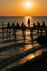 Fototapeta premium Group of friends sit on wooden pier at sunset. Silhouettes of people relax on ocean dock during golden hour. Friends enjoy summer vacation, sea view, calm waves on beach. Peaceful evening lifestyle