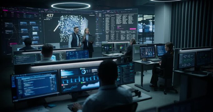 Business Intelligence Manager With a Laptop Presents Ai Neural Network Insights to a Risk Analyst Beside a Huge Particle Display While Colleagues Track Shipping, Banking and Cyber Security Dashboards.