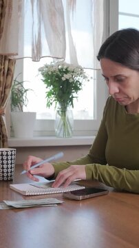 Woman in despair manages family finances, counting cash and writing expenses in notebook as she struggles to make household budget work with too little money, facing financial stress and household