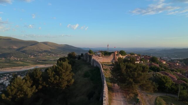Aerial view of the historic stone walls of Berat Castle surrounded by lush trees and rolling hills during golden hour in Berat, Berat County, Albania.