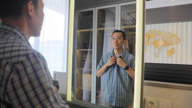 A man looking at his reflection in a mirror while adjusting his shirt in a modern bedroom, with a wardrobe and natural daylight coming through the window. Concept of self-care, personal grooming, dail