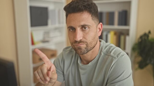 Man with beard in casual shirt points finger at camera beside monitor and bookshelf in studio; serious warning.