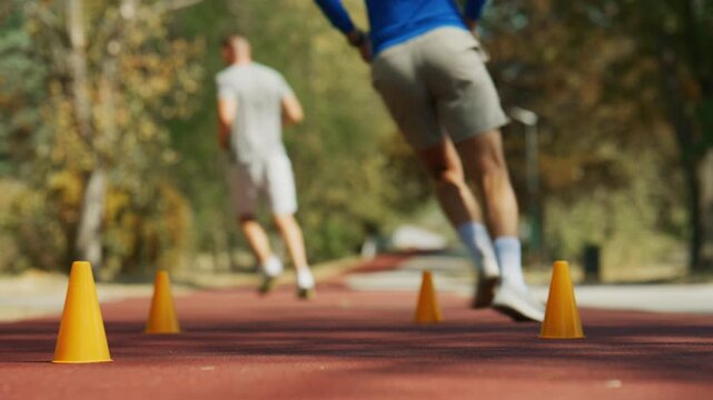 Men perform agility training drills with orange cones on a red running track in a park. Two athletes engage in outdoor fitness and exercise, focusing on speed and coordination.