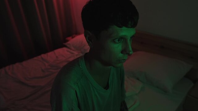 Emotionally withdrawn man sitting alone on his bed, looking down before raising his head and turning away, expressing anxiety, loneliness and inner uncertainty in red and green low key light