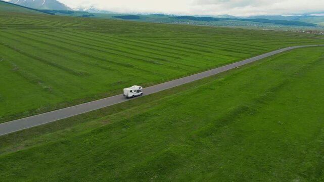 A white camper rides on an empty asphalt road through a vast green landscape with snow-capped mountains in the background, embodying the concept of freedom, travel and adventure vacation in Georgia