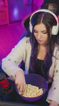 Young woman with long purple hair sits in gaming chair wearing white headphones and reaches for popcorn from large blue bowl. Woman is dressed in fluffy jacket with panda patterns set
