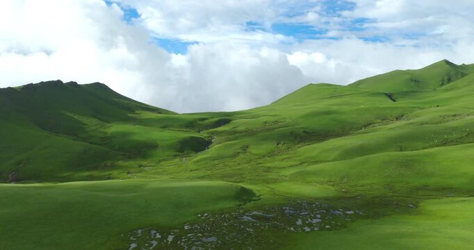 Badimalika region, Bajura, Nepal, aerial shot over lush green alpine pastures, stream beds, and rolling hills in Triveni Patan grassland under blue skies and white cloud formations.