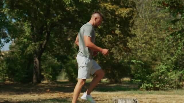 A fit young man is performing step-up exercises on a tree stump in a sunny park. He focuses on his outdoor fitness routine, engaging in a healthy and active lifestyle.
