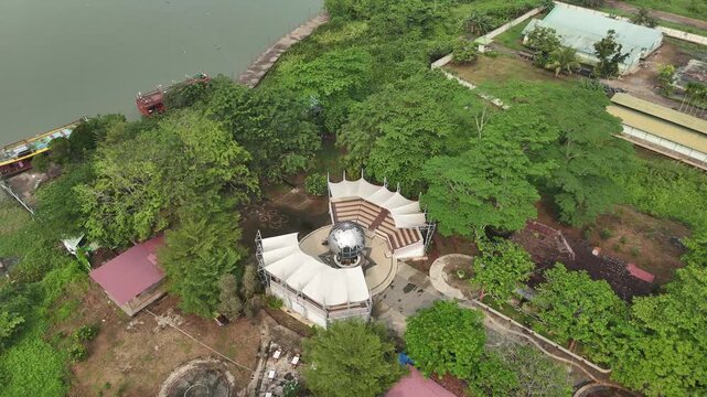 An aerial view of a riverside amphitheater in Pontianak, featuring a white-roofed stage surrounded by lush trees, walking paths, and buildings along the riverbank.