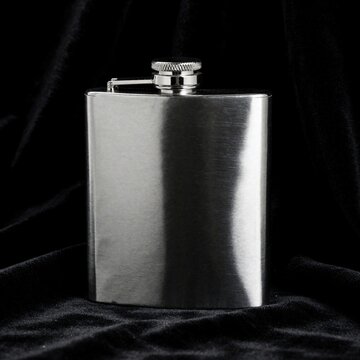 Stainless steel hip flask stands on black velvet fabric. Silver metal container for whiskey vodka sits against dark cloth background. Polished chrome liquor flask used for portable alcohol storage,
