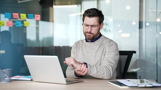 Businessman suffering from wrist pain while working on laptop at desk in office. Male massages wrist joint, performs hand exercises, relieving discomfort, stiffness caused by prolonged computer work.