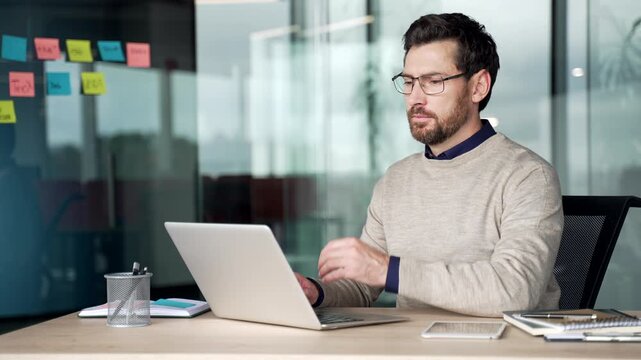 Thoughtful mature businessman working on laptop at desk in office. Pensive professional pauses typing on computer, thinking about business strategy, decisions, focused workday in corporate workplace.