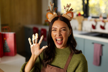 Playful woman wearing Christmas reindeer antlers showing flour-covered hand while baking cookies © Home-stock