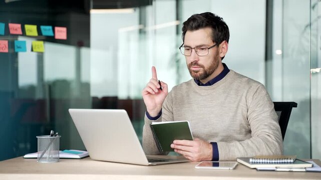 Businessman asking question during online training on laptop writing notes in notebook in modern office. Focused mature professional participating in video conference and remote learning at workplace.