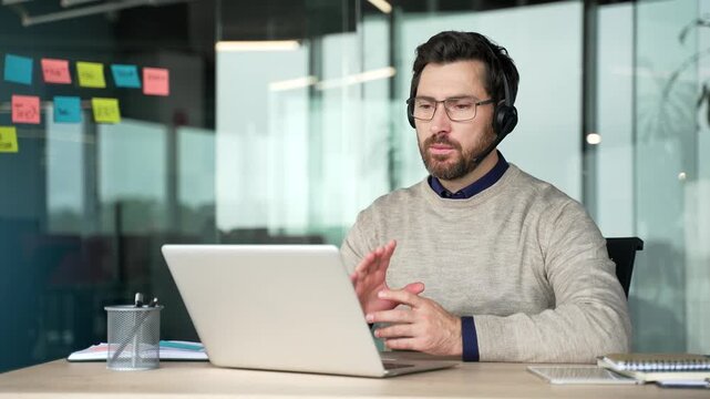 Confident businessman wearing headset conducting online training on laptop in office. Mature professional coach speaking during webinar and communicating with participants in remote learning session.