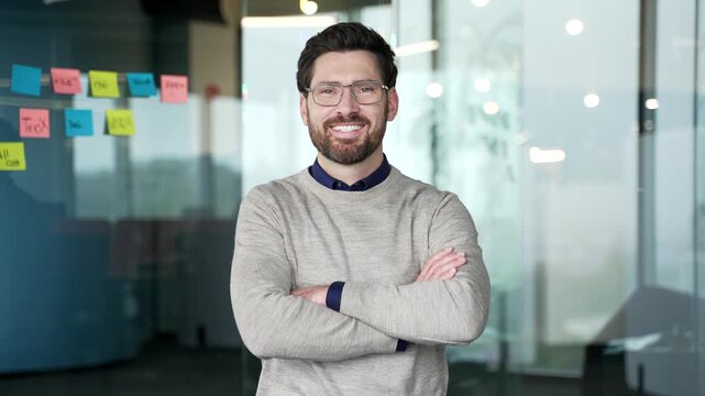 Smiling confident businessman standing with crossed arms in modern glass office. Happy mature professional wearing glasses and sweater looking at camera with positive expression, leadership confidence
