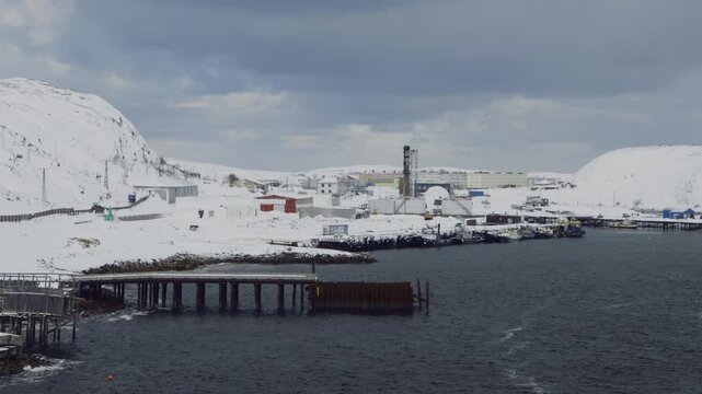 Wide establishing shot of a snowy industrial bay with buildings and docks on the cold coast of the Northern Sea in winter.