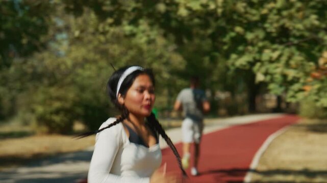 An athletic woman runs on a red track in a sunny park as a man jogs past. The individuals enjoy an invigorating outdoor fitness activity.
