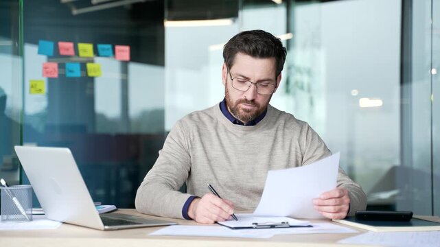 Businessman filling document on clipboard while comparing with paper in hand at desk in modern office. Focused mature professional checking information, verifying data and completing paperwork.
