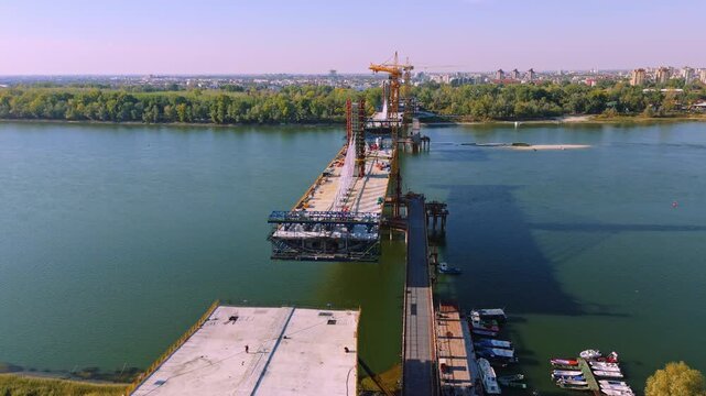 Aerial view of the ongoing bridge construction over the Danube river with concrete sections, construction equipment, and small boats moored along the bank in Novi Sad, Vojvodina, Serbia.