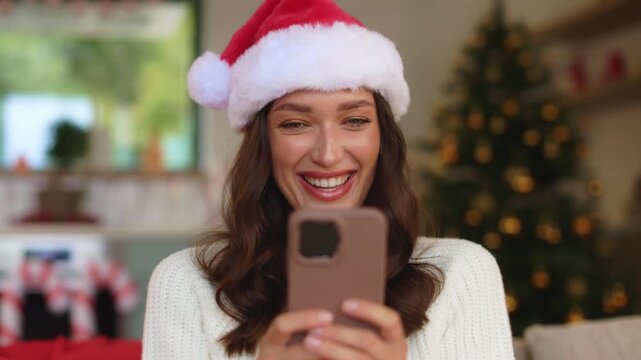 Positive lady in Santa hat looking at cellphone screen and smiling while sitting at home, enjoying pleasant moment during Christmas holiday season, 4K