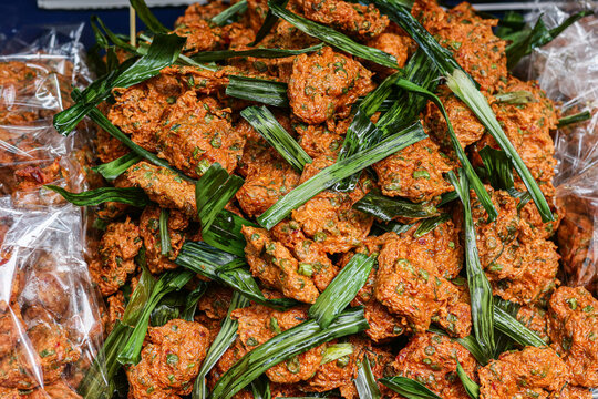 Pile of orange red tod mun fried cakes topped with green pandan leaves, bagged portions at the sides, street food display in Yaowarat, Bangkok, Thailand, full frame close up.