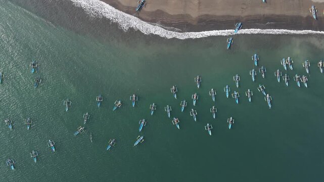 Aerial view of numerous traditional outrigger fishing boats floating in the calm coastal waters near a sandy beach in Cilacap, Central Java, Indonesia.