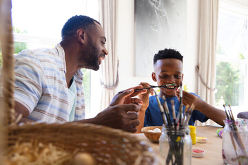 Naklejka premium African American father and youth son painting eggs at dining table by windows with wicker basket