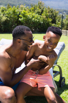 African American father and son shirtless laughing on lounger in garden, trunks drawstring visible
