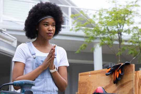 Adult African American woman in denim overalls sanding crate on porch with sander, gloves, earmuffs