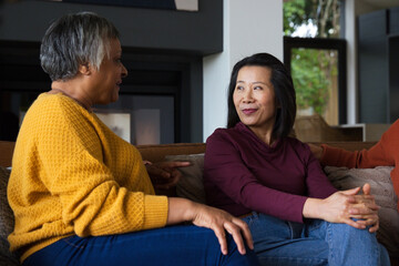 Fototapeta premium Diverse women sitting on sofa, chatting, left gesturing in mustard sweater, right in burgundy top