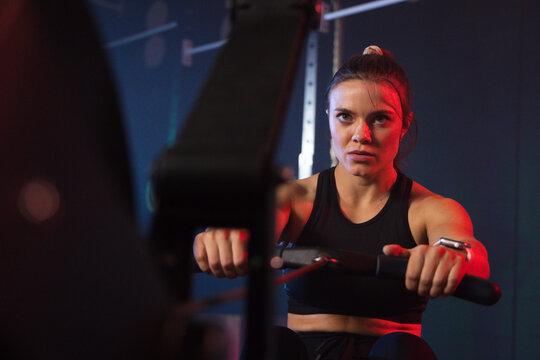 Female rower working on rowing machine in gym, gripping handle, wearing black top and tracker
