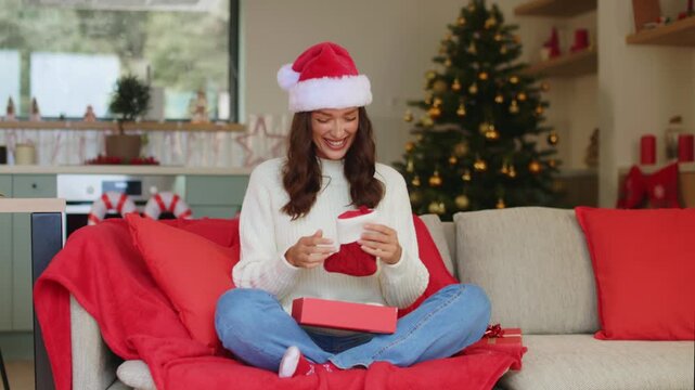 Happy lady in Santa hat receiving gift, opening box and taking out Christmas stockings, smiling happily while enjoying festive holiday moment at home, 4K