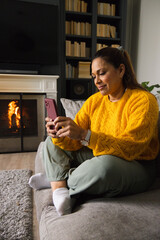 Fototapeta premium African American woman sitting on sofa wearing mustard sweater holding pink phone near fireplace