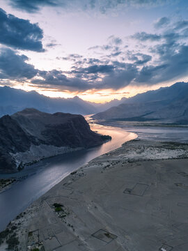Wide aerial landscape showing a meandering Indus River traversing high altitude cold desert sand dunes surrounded by rocky mountain ranges during evening twilight in Skardu Pakistan