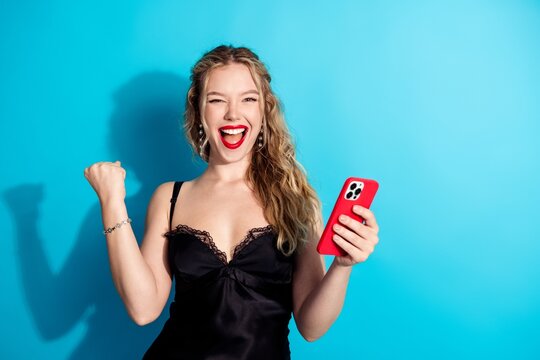 Excited young woman cheering with smartphone in hand, smiling and celebrating a win against a bright blue background