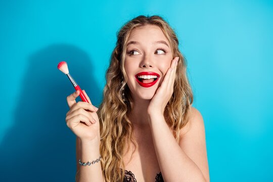 Young woman with red lipstick smiling while holding a makeup brush in a bright beauty portrait
