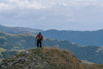 Obraz premium Overweight adult male traveler with a bare torso and large backpack walks alone through high green mountain peaks, moving heavily and persistently while carrying a load, representing endurance