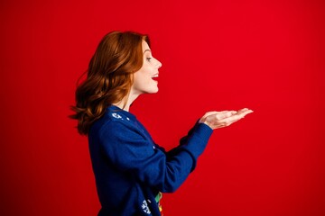 Redheaded woman in blue sweater presenting with open hands against a red background, cheerful profile portrait with holiday sweater vibes and lively expression © deagreez
