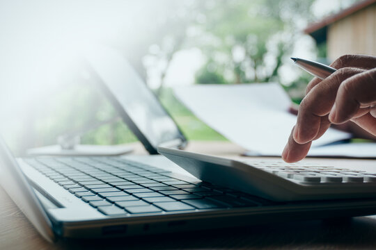 Professional using a calculator with laptop and financial documents in an outdoor natural light setting, concept of business accounting, remote work, and financial calculation.