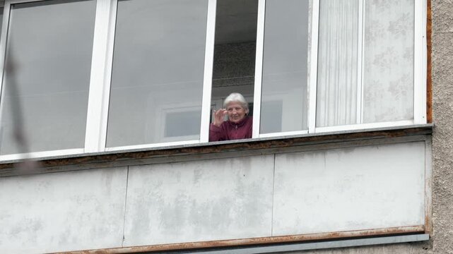 Friendly old grandmother with gray hair standing on balcony of concrete building and waving her hand in greeting, showing a positive and welcoming gesture from apartment window