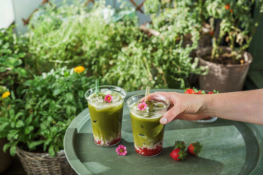 Woman picking up a glass of layered strawberry matcha latte from an outdoor table in a lush green garden setting