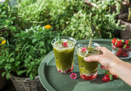 Woman holding a refreshing layered strawberry matcha latte beside another glass on an outdoor table with fresh strawberries and lush greenery.