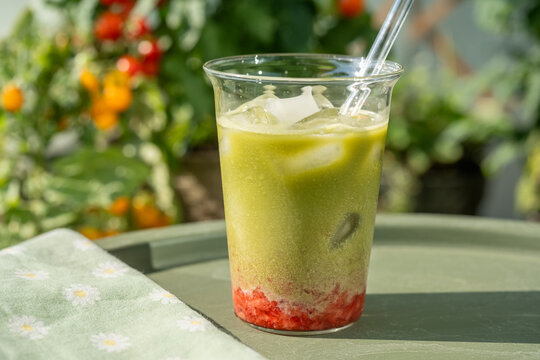 Close-up of a refreshing iced strawberry matcha latte with colorful layers of strawberry, milk and green matcha served outdoors in a summer garden