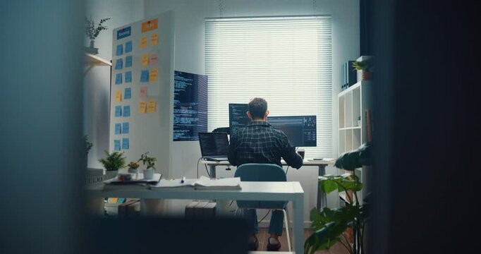 Young Caucasian male programmer writes code using multiple screens in home office workspace. AI development, cybersecurity, network database system, software engineering, remote tech startup concept.