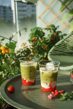 Two refreshing layered strawberry matcha lattes with strawberry, milk and green matcha served outdoors among lush balcony garden plants