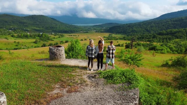 Happy young friends waving and rejoicing on top of a hill as the drone flies away, revealing a beautiful green valley with mountains in Georgia's Racha, a lake and a village road on a sunny day with c