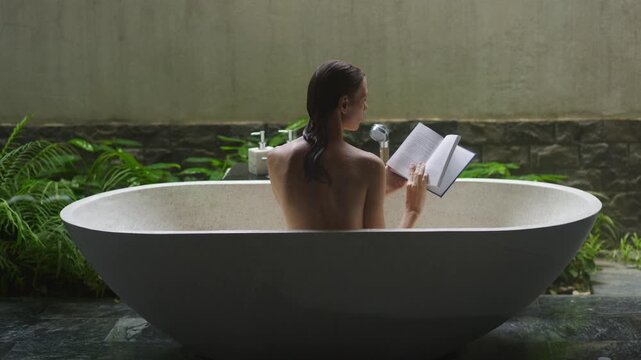 Woman reading book while sitting in stone bathtub in bright balinese style bathroom