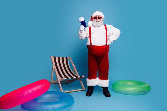 Santa Claus on summer vacation holding passport and boarding pass with inflatable rings and deck chair against bright blue background